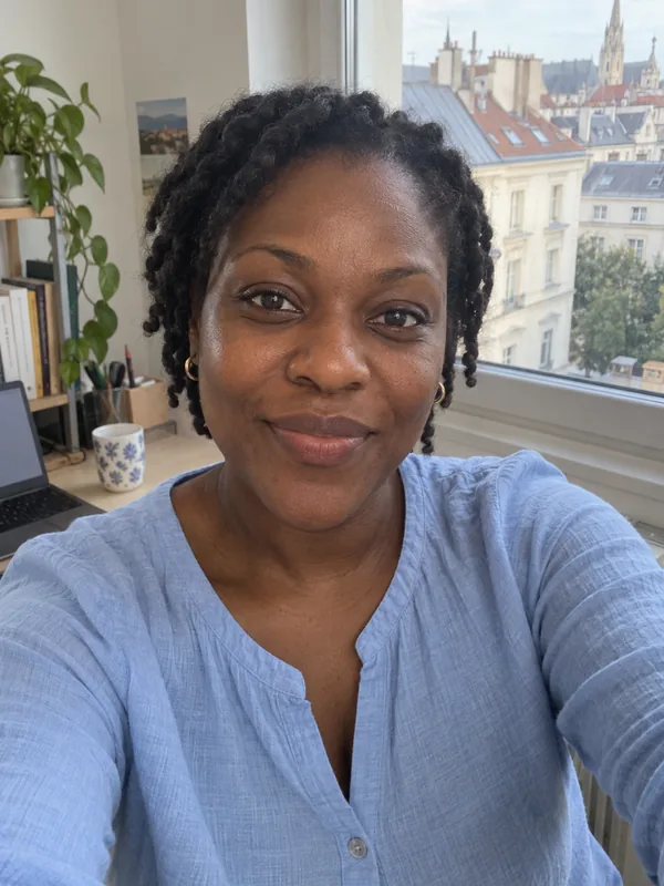 Handy-Selfie einer Frau um die 35 mit kurzen Twists und hellblauer Bluse am Schreibtisch, hinter ihr die Dächer der Stadt.