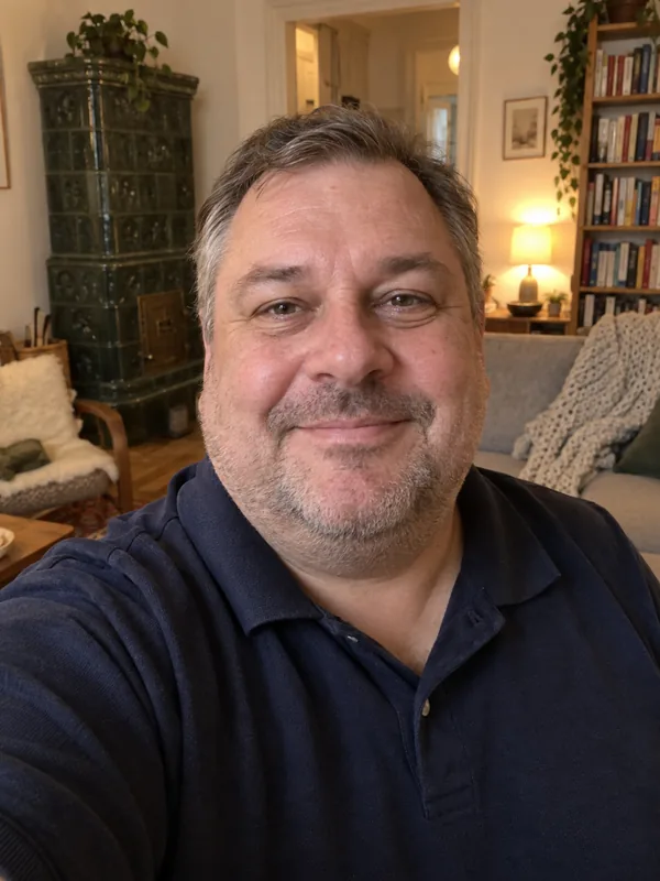 Handy-Selfie eines kräftigen Mannes um die 50 mit graumeliertem Haar und Bartstoppeln im Wohnzimmer mit Kachelofen.