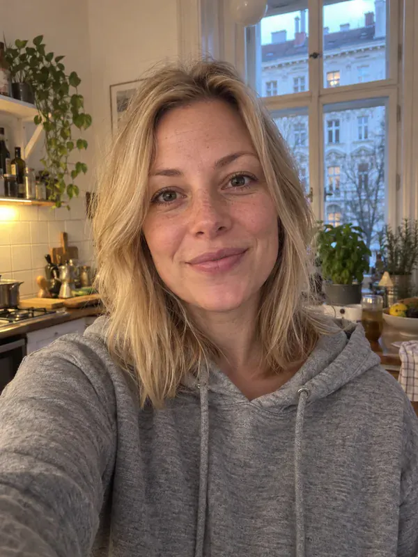 Handy-Selfie einer Frau um die 30 mit schulterlangen blonden Haaren und grauem Kapuzenpulli in einer Altbauküche.