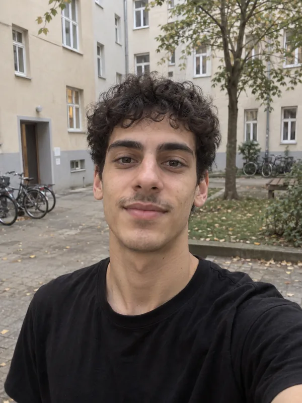 Handy-Selfie eines schlanken jungen Mannes mit dunklen Locken und schwarzem T-Shirt in einem Wohnhof.